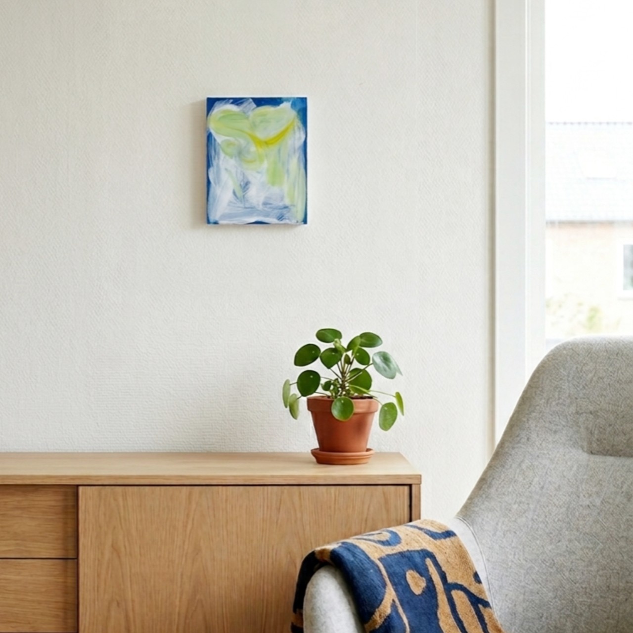 OtonoSasayaki-series02/片岡由香里(カタオカ・ユカリ)による、青、白、黄色の筆致が特徴的な抽象画が、木製のキャビネットと鉢植えの上、模様入りのスローがかけられたアームチェアの近くに飾られている。.