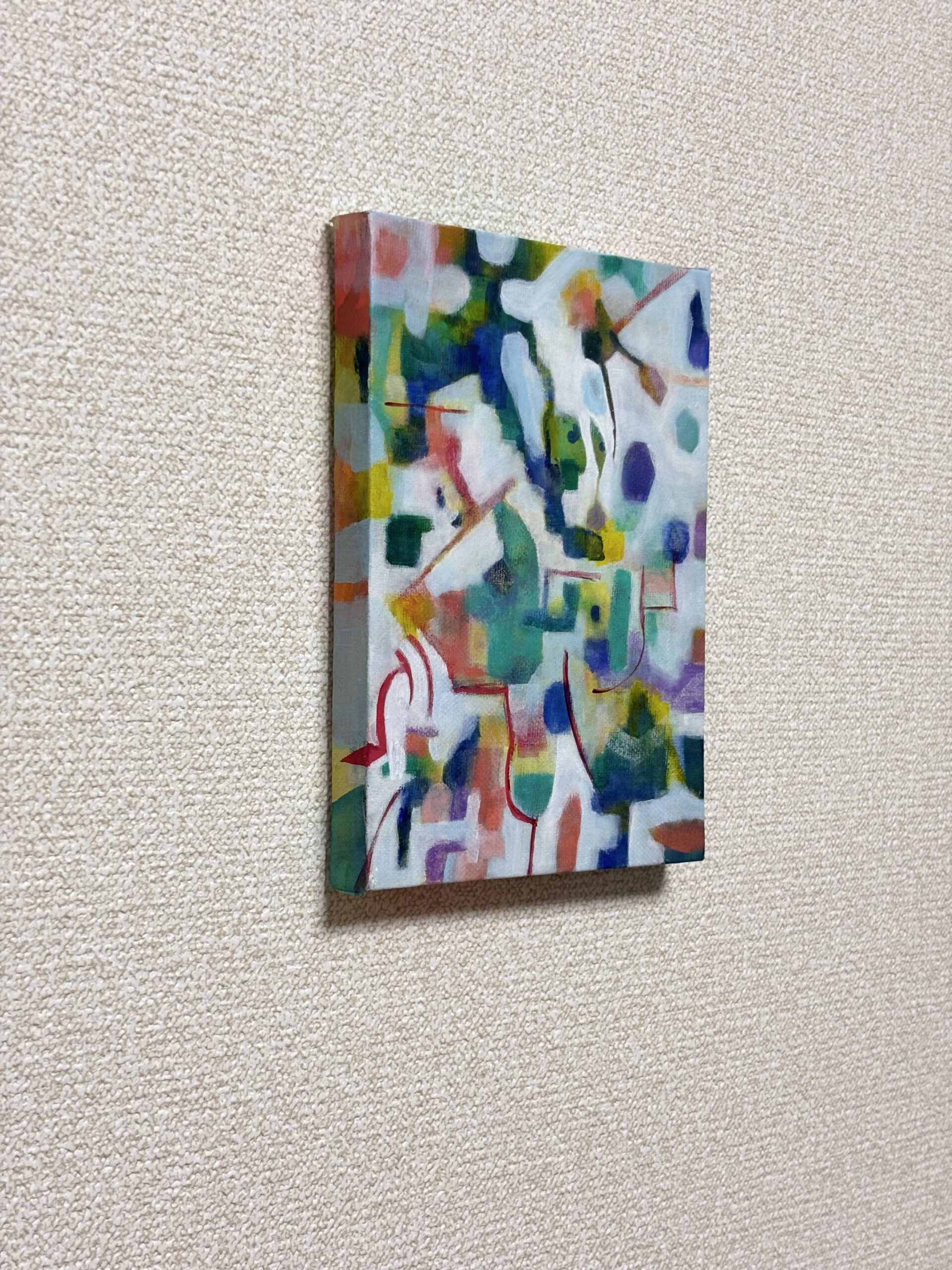 接続の試行と題された、知念由里(チネン・ユリ)による小さな長方形の抽象画。青、緑、黄色、赤のカラフルな形や線が描かれており、質感のある白い壁に掛けられている。.